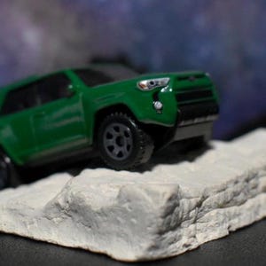 Peut inclure: Un SUV jouet vert est positionné sur une petite formation rocheuse grise texturée. Le véhicule a des roues noires et une calandre noire. L'arrière-plan est un mélange flou de bleu, de violet et de blanc.