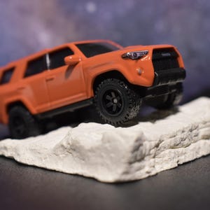 May include: A bright orange die-cast Toyota SUV toy vehicle featuring black wheels, a black front grille, and detailed headlights. The miniature truck is positioned at an angle on a textured, off-white rock-like base, showcasing its rugged design.
