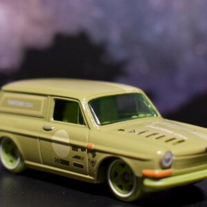 May include: A light olive green die-cast station wagon toy car featuring white circular decals on the doors, white racing stripes on the hood, and the text Pantone 2334 printed on the side panel and hood. The vehicle has green tinted windows.