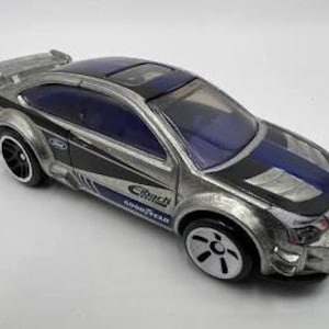 Könnte beinhalten: Ein silbernes und schwarzes Spielzeugauto mit blauem Dach und Akzenten. Das Auto hat das Ford-Logo und das Branding von Goodyear und Eibach. Die Räder sind schwarz und weiß.