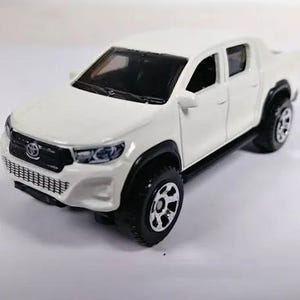 Könnte beinhalten: Ein weißes Spielzeug-Pick-up mit schwarzen Rädern und Akzenten. Der Truck hat einen detaillierten Frontgrill, Scheinwerfer und ein Toyota-Emblem. Die Reifen sind schwarz mit silbernen Felgen. Ein Miniatur-Replikat.