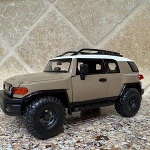 May include: A tan and white toy SUV with black tires and a black roof rack. The vehicle has a black front bumper, side mirrors, and door handles. The headlights are orange, and the taillights are red.