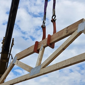 May include: A wooden roof truss being lifted by a crane. The truss is made of light-colored lumber and has metal connector plates. The crane uses purple straps and orange lifting clamps to secure the truss. The background is a blue sky with white clouds.