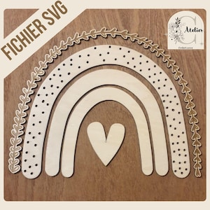 May include: Wooden rainbow SVG design with a heart in the center. The rainbow has a dotted pattern and is surrounded by a leaf design. The text "FICHIER SVG" is in the upper left corner. The logo for "Atelier Créations" is in the upper right corner.