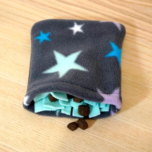 Könnte beinhalten: Ein dunkelgraues Fleece-Haustierspielzeug mit Sternenmuster. Das Spielzeug ist oben offen und zeigt einen hellblauen Innenraum mit braunen Leckerlis. Mehrere Leckerlis liegen auf einer Holzoberfläche verstreut.