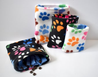 Op de afbeelding: Een verzameling fleece traktatiezakjes voor huisdieren in verschillende maten en kleuren. De zakjes hebben een pootafdrukpatroon in blauw, oranje, roze en bruin op een witte of zwarte achtergrond. Sommige zakjes zijn open en tonen blauwe stof en traktaties.