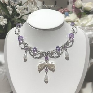 May include: A silver chain necklace with purple and white beads, a white bow charm, and pearl drop accents. The necklace is on a white mannequin.