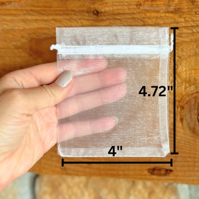 May include: A sheer white organza gift bag, 4 inches wide and 4.72 inches tall, with a white drawstring closure. The bag is held against a wooden background, showcasing its translucent quality.