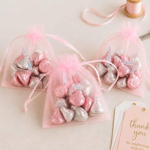 May include: Three sheer pink organza drawstring bags filled with silver and pink foil-wrapped chocolate candies. Each bag features a small white ribbon tag with the word Kisses printed on it. A pink gift tag with the text thank you for celebrating with us is visible.