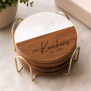 May include: A stack of circular coasters featuring a split design of white marble and dark brown wood, held in a gold metal wire stand. The wood portion is engraved with the text THE Keichens EST. 2013 in a dark, script-style font.