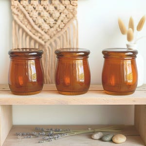 May include: Three amber glass jars with matching lids, each featuring a rounded, wide-body design. The jars are made of translucent brown glass, providing a clear view of the interior, and are topped with smooth, circular glass lids.