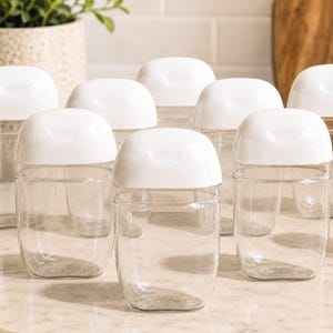 May include: A collection of eight clear plastic containers with rounded white flip-top lids. Each container features a smooth, transparent body and a secure, hinged cap design, arranged in a group to display their uniform shape and size.