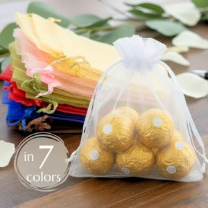 May include: A white sheer organza drawstring bag filled with five gold foil-wrapped round candies. Behind it, a stack of similar bags in yellow, pink, green, red, and blue is visible. A circular graphic overlay displays the text in 7 colors.