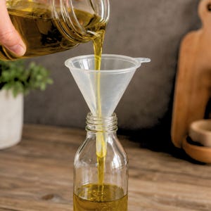 May include: A clear plastic funnel is positioned in the neck of a transparent glass bottle. Golden yellow liquid is being poured from a glass container through the funnel into the bottle, filling the bottom portion with the translucent liquid.