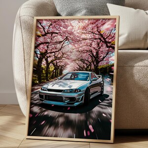 Könnte beinhalten: Gerahmter Kunstdruck mit einem silbernen Sportwagen, der eine von Kirschblütenbäumen gesäumte Straße entlangfährt. Das Bild hat einen Bewegungsunschärfe-Effekt, mit fallenden rosa Blütenblättern. Das Kunstwerk wird auf einem Holzboden ausgestellt.
