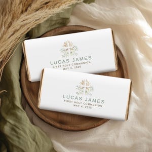 Puede incluir: Dos envoltorios de barra de chocolate blanco con una ilustración de acuarela de una cruz con vegetación y el texto "Lucas James Primera Comunión 4 de mayo de 2025".