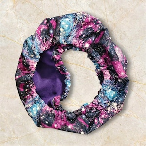 May include: A circular hair bonnet featuring a galaxy design in shades of purple, blue, and white on a black backdrop. The interior is a solid purple, and the bonnet has an elasticated edge.
