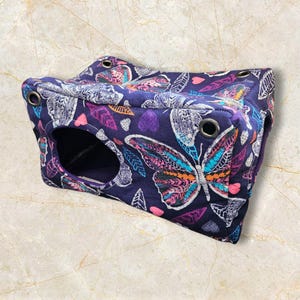 May include: A navy blue pet bed featuring a butterfly and leaf design. The bed is rectangular with a circular entrance and metal eyelets. The butterfly wings are in shades of orange, teal, and pink.