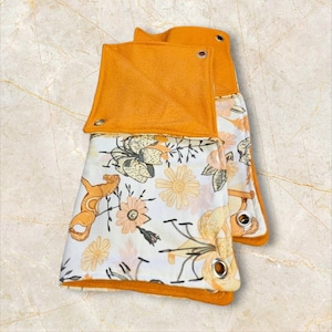 May include: Two folded blankets with a floral pattern and orange trim. The blankets feature a white background with a pattern of flowers in shades of yellow, orange, and brown. The trim is a solid orange color, and each blanket has metal grommets.