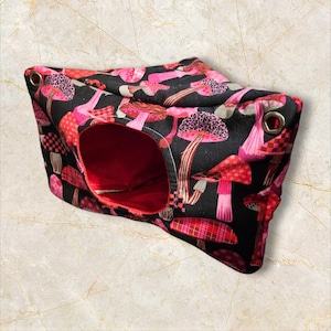 May include: A small pet hideaway with a black exterior covered in a pink and red mushroom pattern. The interior is a solid red colour. The hideaway has a circular opening and metal eyelets on the corners.