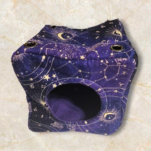 May include: A purple pet bed with a celestial design featuring constellations, moons, and stars. The bed has a circular opening and is made of soft fabric. The bed has three metal eyelets on the top.