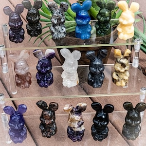 Puede incluir: Una colección de figuras de conejos de piedra tallada en varios colores, incluyendo morado, negro, azul y crema. Los conejos se exhiben en un soporte transparente de varios niveles, mostrando su detallada artesanía y texturas únicas. El fondo es un entorno natural con vegetación.