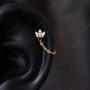 May include: A gold ear piercing with a three-stone cluster design and a dangling chain. The cluster features clear, sparkling stones. The chain connects to a second piercing. The image is set against a dark background.