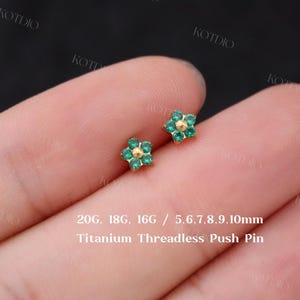 May include: Two small, gold-coloured flower-shaped earrings with green and gold accents. The earrings are shown on a hand. Text on the image reads "20G. 18G. 16G / 5.6.7.8.9.10mm Titanium Threadless Push Pin."