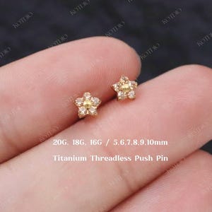 May include: A pair of gold-coloured star-shaped earrings, each with a central yellow stone and surrounding clear stones. The earrings are small and delicate, with the text "Titanium Threadless Push Pin" and size information.