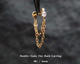 Puede incluir: Un pendiente de cadena doble con parte trasera plana de color dorado. El pendiente presenta una pequeña gema redonda y transparente y un diseño de cadena colgante. El texto "Double Chain Flat Back Earring 18G / 6mm" es visible.