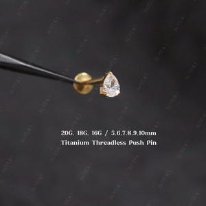 Könnte beinhalten: Ein goldfarbener Titan-Ohrstecker ohne Gewinde mit einem klaren, tropfenförmigen Edelstein. Der Ohrring wird von einem schwarzen Werkzeug gehalten. Text auf dem Bild: 20G, 18G, 16G / 5.6,7,8,9,10mm Titanium Threadless Push Pin.