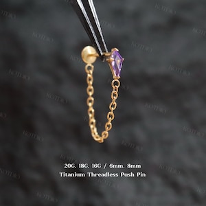 Könnte beinhalten: Ein goldfarbener Titan-Ohrstecker ohne Gewinde mit Kette und lila Edelstein. Der Ohrring wird mit einer Pinzette vor dunklem Hintergrund gehalten. Der Text "Copyright @kotdio" und "20G, 18G, 16G / 6mm, 8mm Titanium Threadless Push Pin" sind sichtbar.
