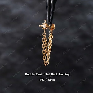May include: A gold-coloured double chain flat back earring. The earring is held by tweezers. The earring has a star-shaped top with a small gem and two dangling chains. The text "Double Chain Flat Back Earring" and "18G / 6mm" is visible.