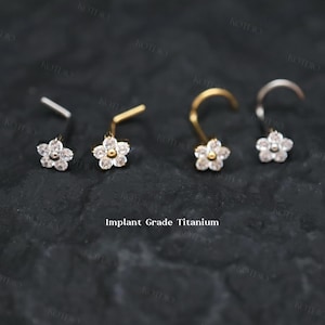 May include: Four nose studs with flower-shaped designs. Two are straight, and two are curved. Each flower has a central gem surrounded by smaller gems. The text "Implant Grade Titanium" is visible.