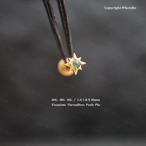 May include: A gold-toned starburst-shaped body jewelry stud featuring a central round green stone. The text reads 20G, 18G, 16G / 5, 6, 7, 8, 9, 10mm Titanium Threadless Push Pin. The piece is held by dark tweezers against a dark background.