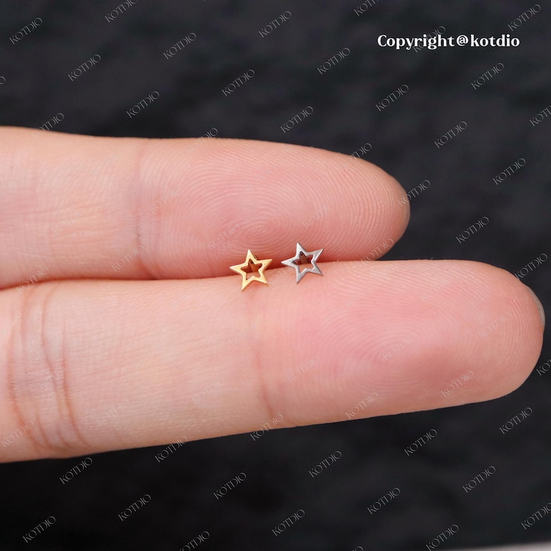 Op de afbeelding: Twee stervormige oorringen, een gouden en een zilveren, worden getoond op een vinger. De oorringen zijn klein en delicaat, met een hol centrum.