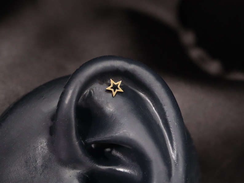 Op de afbeelding: Een gouden stervormige oorbel met een zwarte omtrek. De oorbel wordt in close-up weergegeven op een zwarte achtergrond.