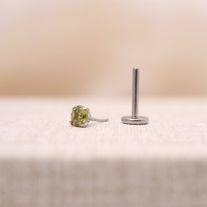 May include: A silver body jewelry stud with a green gemstone. The stud is shown on a white fabric background.