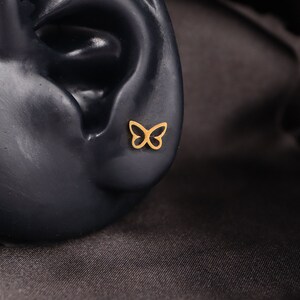 May include: A gold butterfly earring stud. The earring is shown on a black mannequin ear.