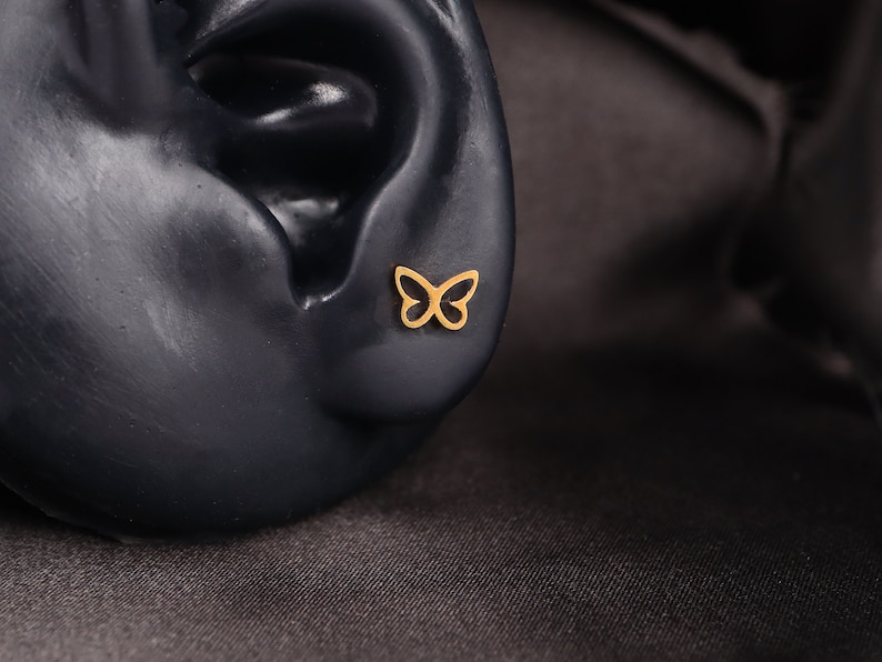 May include: A gold butterfly earring stud. The earring is shown on a black mannequin ear.