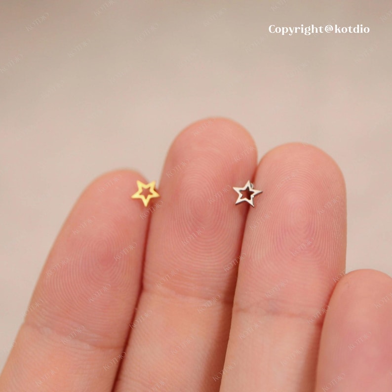 Op de afbeelding: Twee kleine stervormige oorbellen, een gouden en een zilveren, worden getoond op een vinger. De oorbellen zijn hol en hebben een eenvoudig ontwerp.