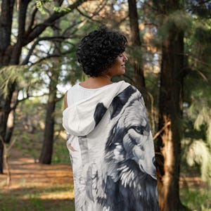 May include: A hooded blanket featuring a detailed grayscale wolf portrait. The blanket is white with a black and gray wolf design. The person is wearing a white tank top. The background is a forest with tall trees.