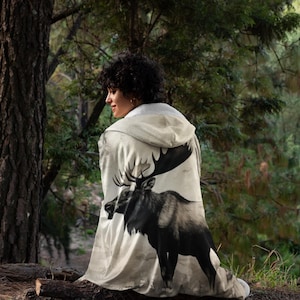 Puede incluir: Una manta con capucha de color crema con una ilustración en blanco y negro de un alce. La manta se usa al aire libre en un bosque. La imagen del alce es detallada y cubre la parte posterior de la manta, con una capucha.