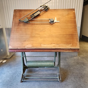 May include: An antique drafting table with a wooden top and a metal base. The table features a drawing arm and a roll holder. The wooden top is approximately 122 cm wide and 91 cm deep. The metal base is painted green.