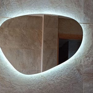 Peut inclure: Intérieur de salle de bain moderne avec un lavabo en forme de vasque à motifs marbrés et un robinet chromé. Au-dessus, un miroir ovale avec éclairage intégré est fixé sur un mur texturé en imitation pierre.