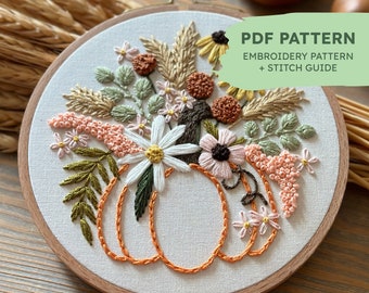 May include: Embroidered hoop art featuring a pumpkin with fall floral accents. The design includes a variety of flowers, leaves, and stems in shades of pink, green, brown, and white. The text "PDF PATTERN EMBROIDERY PATTERN + STITCH GUIDE" is visible in the upper right corner.