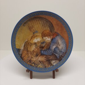 Puede incluir: Un plato de cerámica azul y dorado con una pintura de una pareja abrazándose frente a un arpa. El plato está en un soporte de madera.