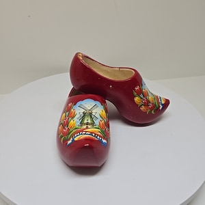 May include: A pair of red wooden clogs with a windmill and tulip design. The clogs are painted with a blue sky, green grass, and yellow tulips. The text "HOLLAND" is written on a blue ribbon.