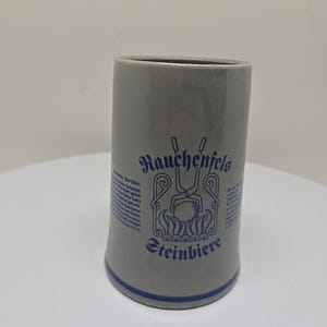 May include: A gray ceramic beer stein with a blue and white design. The stein features the text "Rauchenfels Steinbiere" and a graphic of a blacksmith's tongs and a ball of metal.