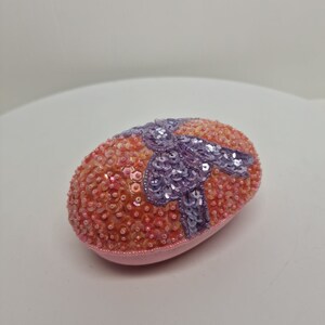 May include: An Easter egg-shaped ornament covered in orange and pink sequins. A lavender sequined bow is centered on the egg. The base of the egg is a soft pink fabric.